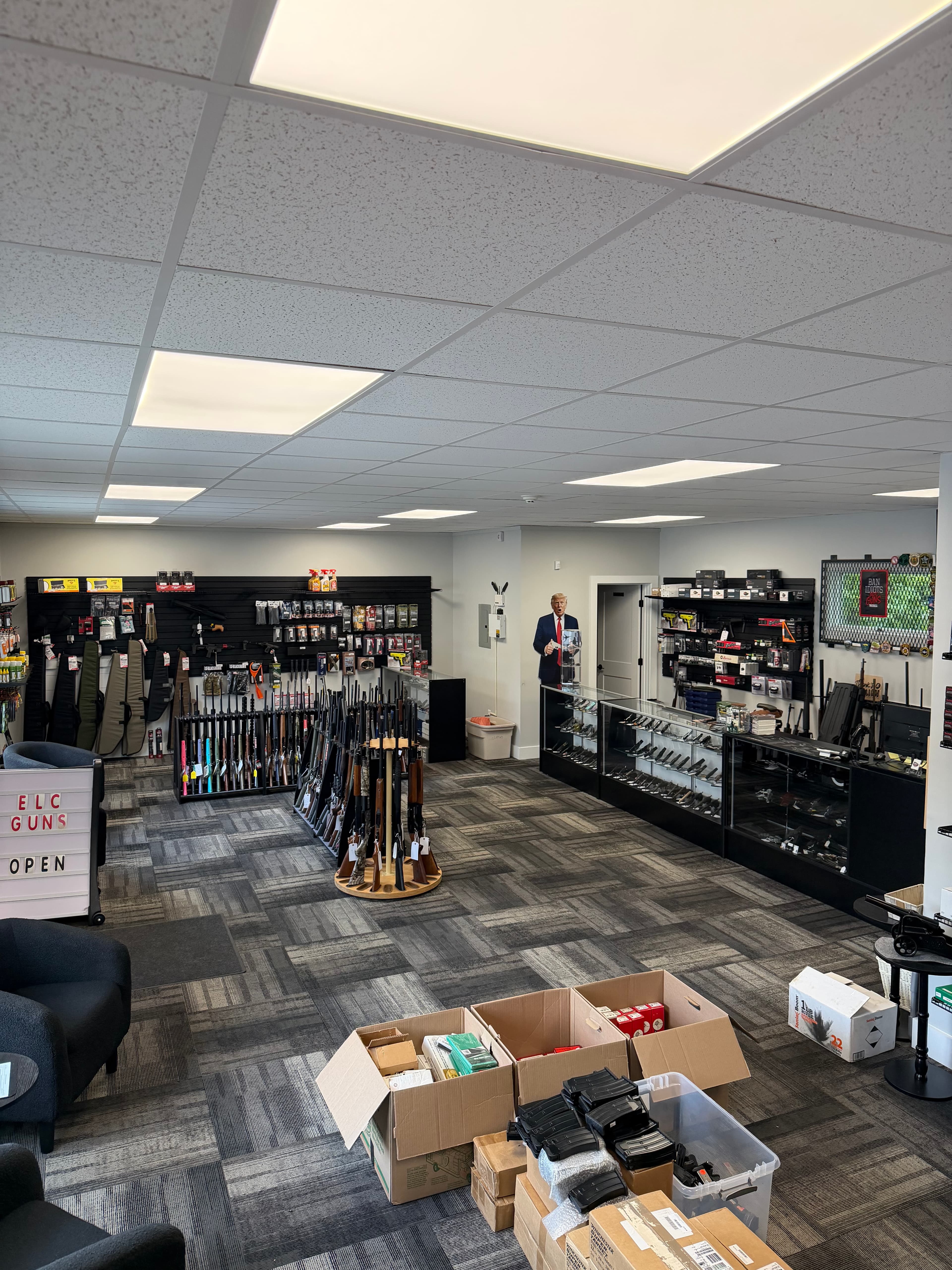 Gun shop interior with rifles on racks, handguns in cases, and a Trump cutout.