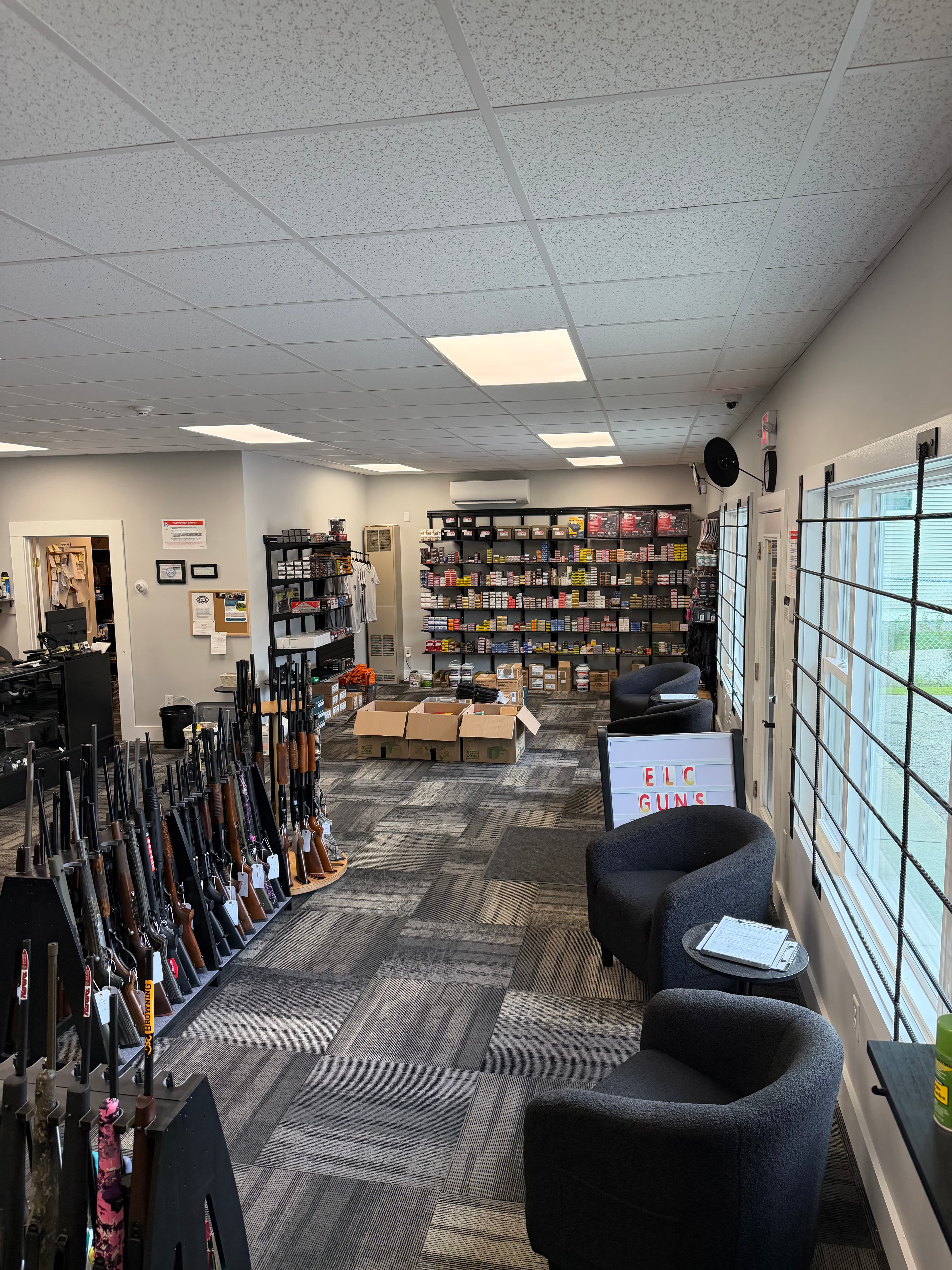 Gun shop interior featuring rifle display racks, ammunition shelves, and grey armchairs for customers.